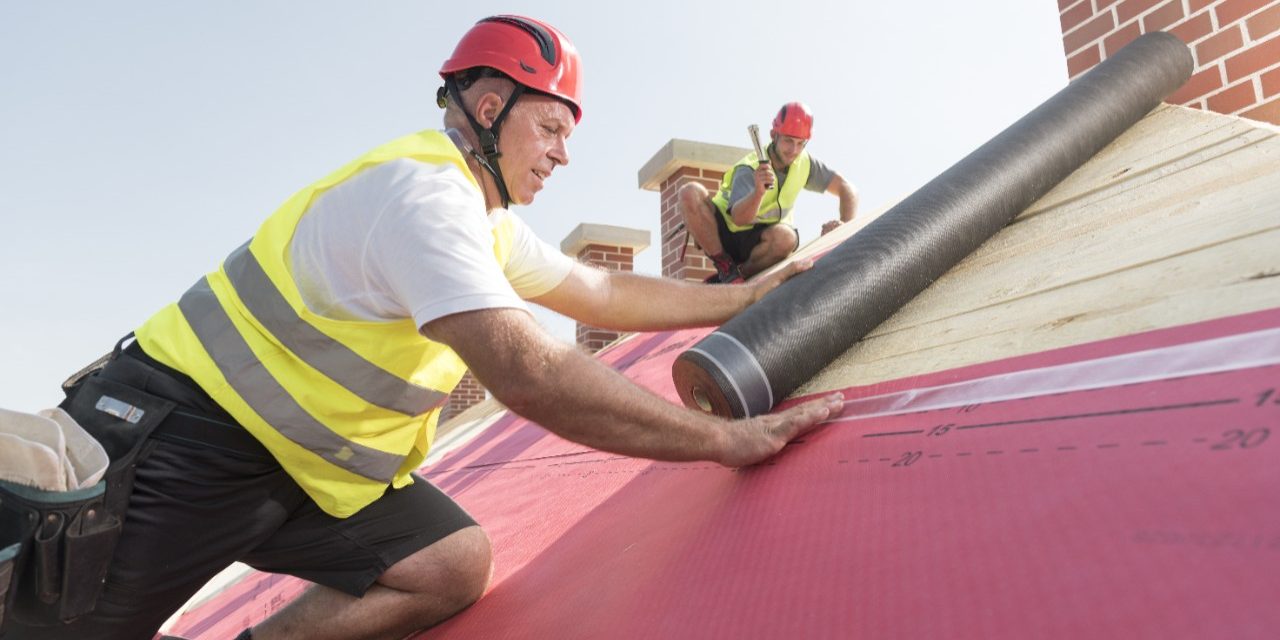 Urban roofers applying roof underlay sheet wearing hard hat and safety jacket brick chimney