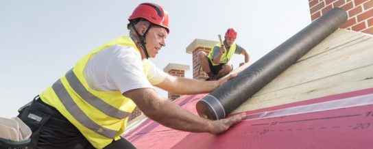 Urban roofers applying roof underlay sheet wearing hard hat and safety jacket brick chimney