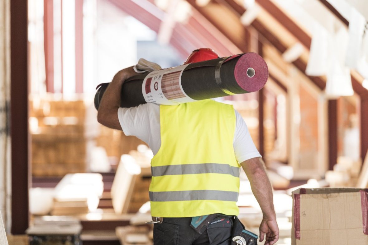 Urban roofer carrying underlay sheet on his shoulder in renovated attic wearing hard hat and safety jacket